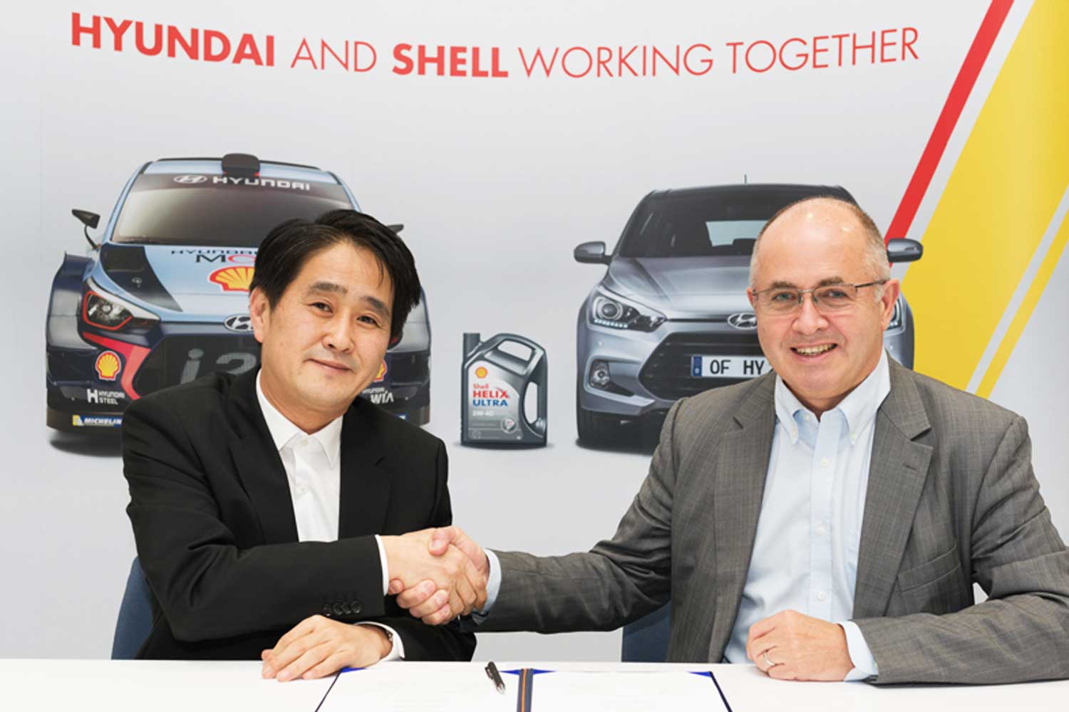 Representantes de Hyundai y Shell estrechando manos durante la firma de un acuerdo de colaboracion con vehiculos y lubricantes Shell Helix Ultra de fondo